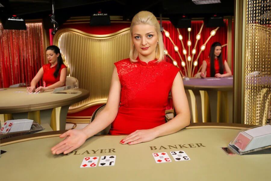 Bakarat na żywo w kasynie online. blondyna w czerwonym prowadzi grę i rozdaje.